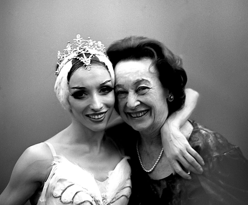 Natalia Makrova and Lucia Chase are pictured from the chest up, thei cheeks pressed together as they smile towards the camera. Makarova is wearing a white swan tutu and a tiara with feathers alongside both ears, and she wraps her left arm around Chase, who is wearing a pearl necklace and a dark top.