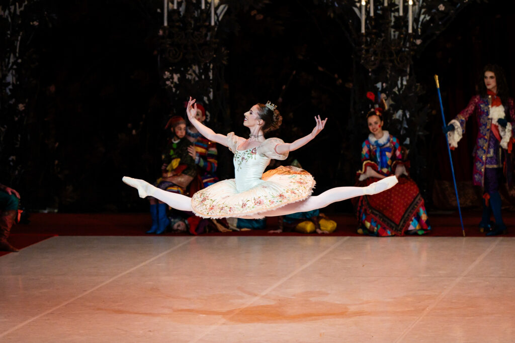 Abigail Willson-Heisel splits her legs in a saut de chat across the stage, arms curved up gently to either side. She smiles, wearing a light cream and peach-colored tutu with puffy short sleeves and rose ornamentation. Behind her, courtiers sit and watch.