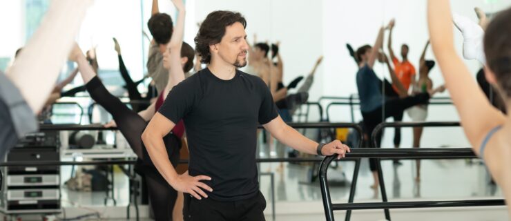 Cervilio Miguel Amador, wearing a black t-shirt and black pants, is shown from the knees up with his right hand on his hip as he rests his left hand on the end of a portable ballet barre. He looks to his left, watching dancers as they practice grand battement during a ballet class.