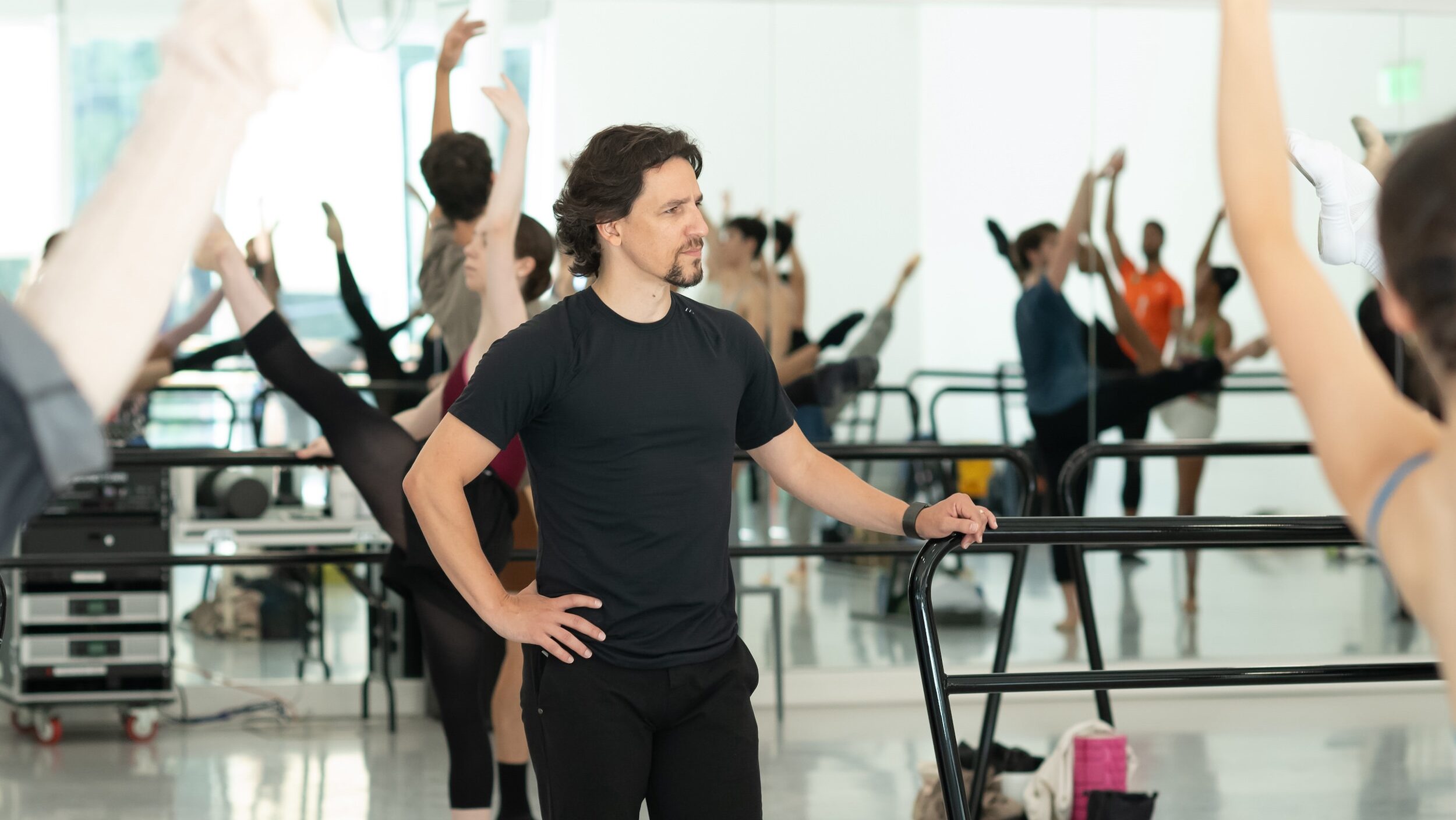 Cervilio Miguel Amador, wearing a black t-shirt and black pants, is shown from the knees up with his right hand on his hip as he rests his left hand on the end of a portable ballet barre. He looks to his left, watching dancers as they practice grand battement during a ballet class.