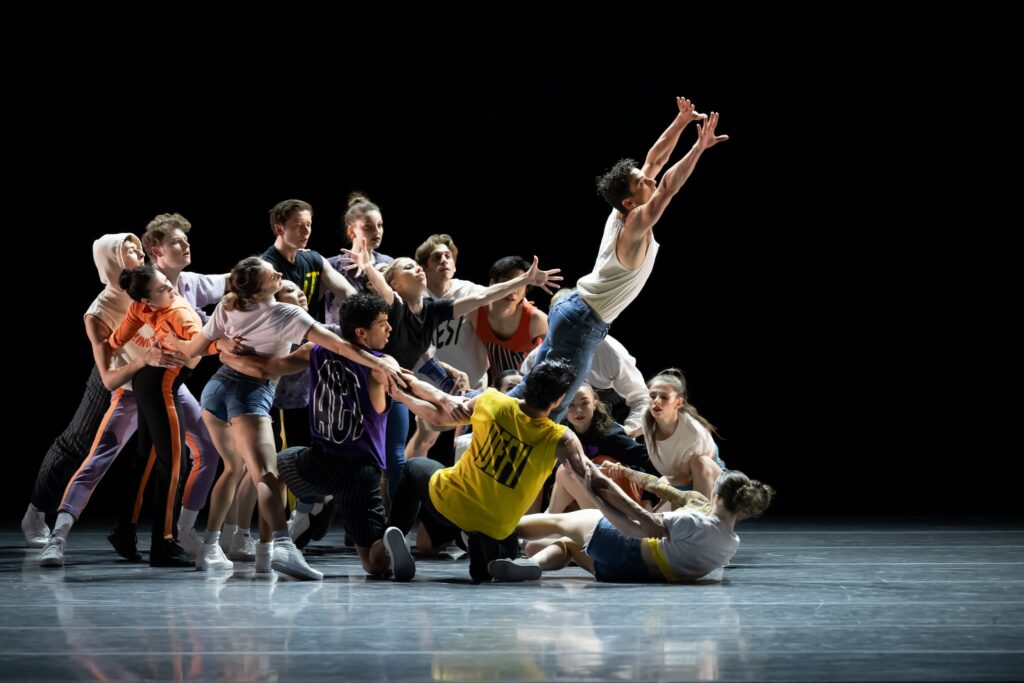 A large corps of dancers, dressedin casual street wear, perform onstage in front of a dark backdrop. The group forms a circular clump, linking hands and arms, with the dancers gradually lowering the formation to the floor towards the left. A lone male dancer in a white t-shirt and jeans jumps up energentically from the circle's center, his arms reaching high and his body projecting out diagonally towards stage left.