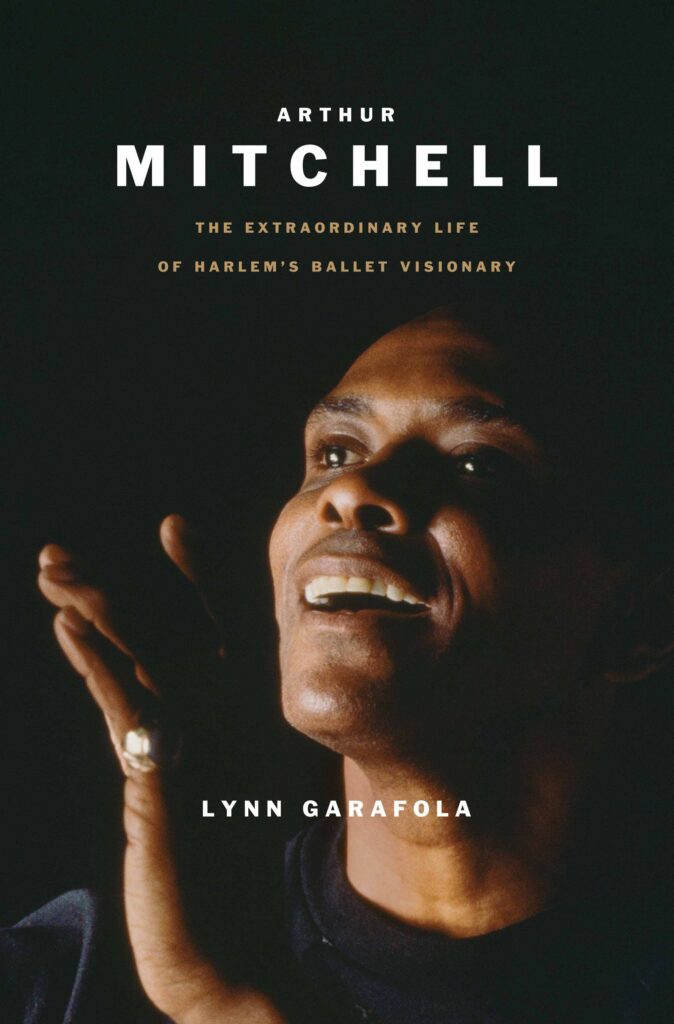The cover of a biography titled "Arthur Mitchell: The Extraordinary Life of Harlem’s Ballet Visionary." On the cover is a photograph of Arthur Mitchell lit from the side against a dark background. He opens his mouth to speak with a pleasant expression, one hand raised with fingers gently bent.