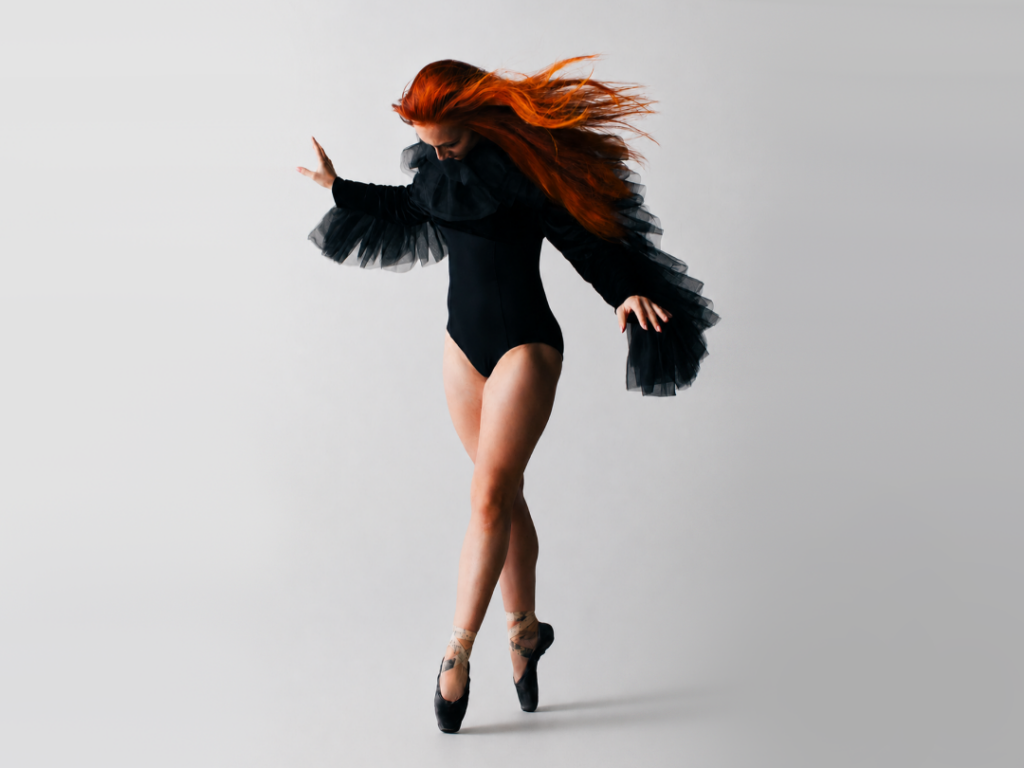 Megan Boyette is shown against a white backdrop in croisé fourth position en pointe. She wears black pointe shoes and a black leotard with long-sleeves lined with wing-like tulle that also gathers at her neck. Boyette looks down at her feet, her long red hair blowing away from her face.