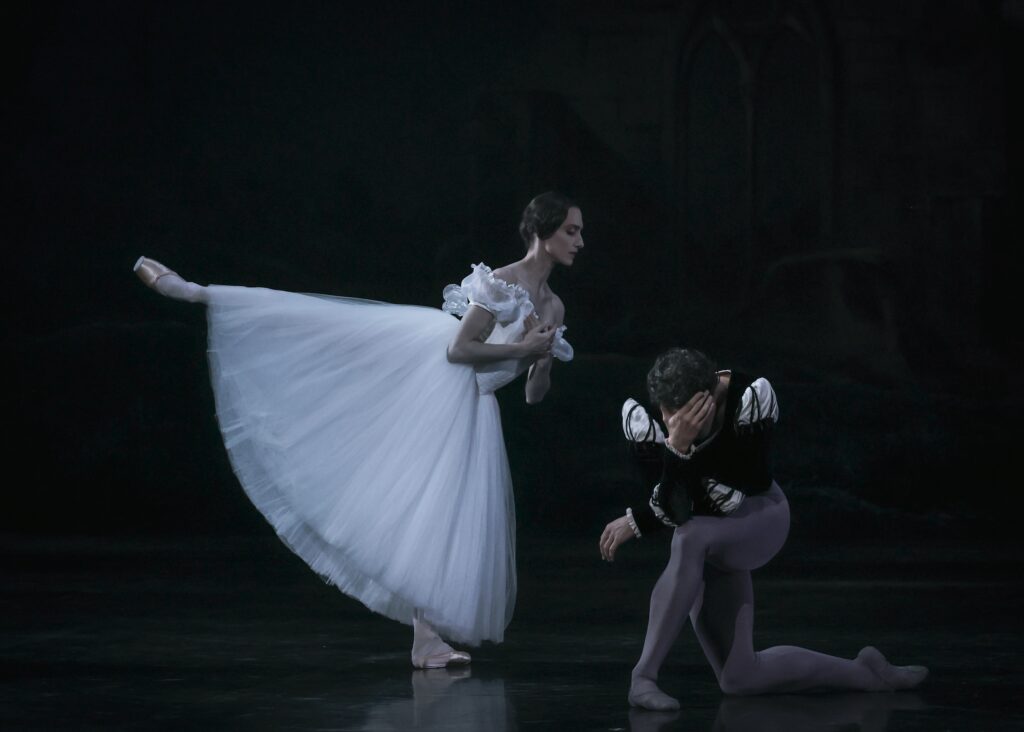 Dorothée Gilbert, wearing a white Romantic tutu and pointe shoes, does an arabesque towards her partner, Hugo Marchand, pressing her hands to her heart. Marchand, wearing a black velvet tunic and gray tights, kneels beside her and covers his face with his right hand in despair.