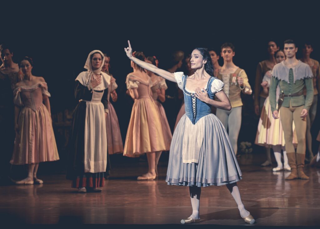 Dorothée Gilbert, wearing a blue peasant dress, tights, and pointe shoes, stands on her right leg, her left slightly out to the side. Her dark hair is down as she clutches her heart with her left hand and points her right index finger out in front of her. Other dancers costumed as peasant villagers stand behind her and watch with concerned expressions.