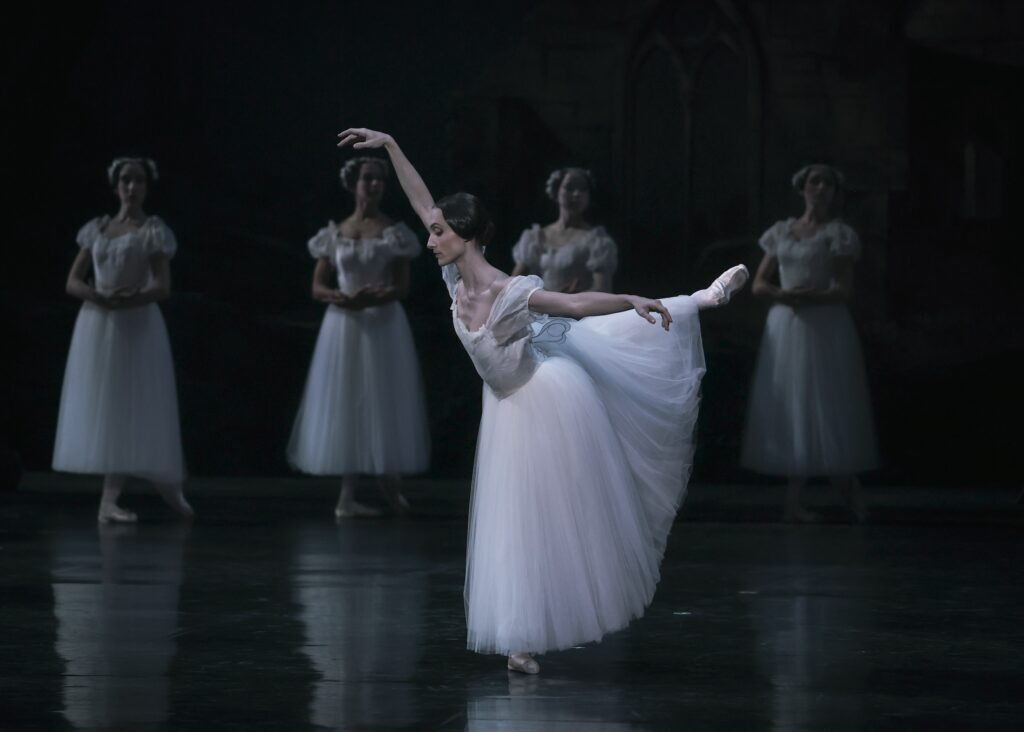 Dorothée Gilbert is shown in a third arabesque allongé and wears a white Romantic tutu and pointe shoes. A line of ballerinas in similar costumes stand in B plus with their wrists crossed in a line in the background.