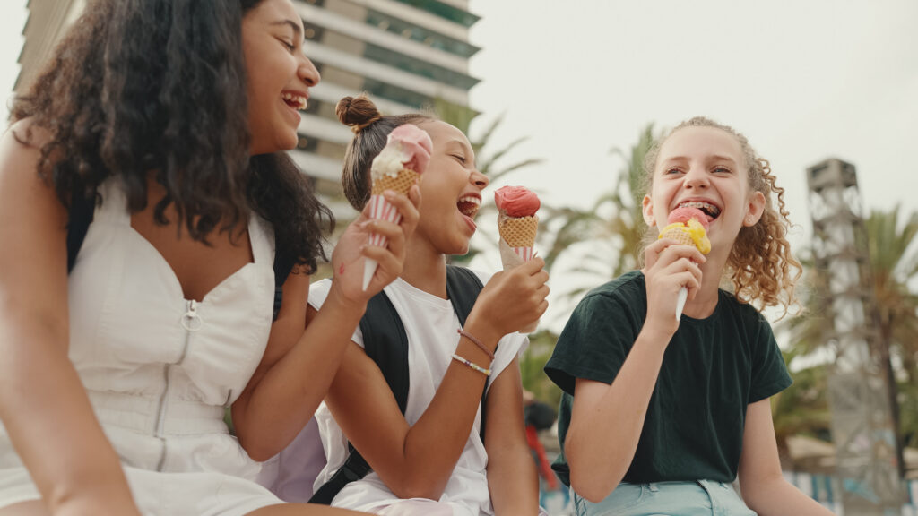 Three young teenagers sit together outside laughing and holding ice cream cones.