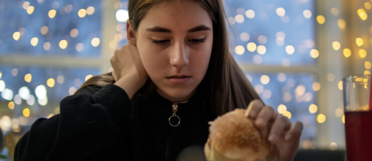 A teenage girl sits at a table with one hand on her neck, holding a cheeseburger on her plate. She looks at it reluctantly.