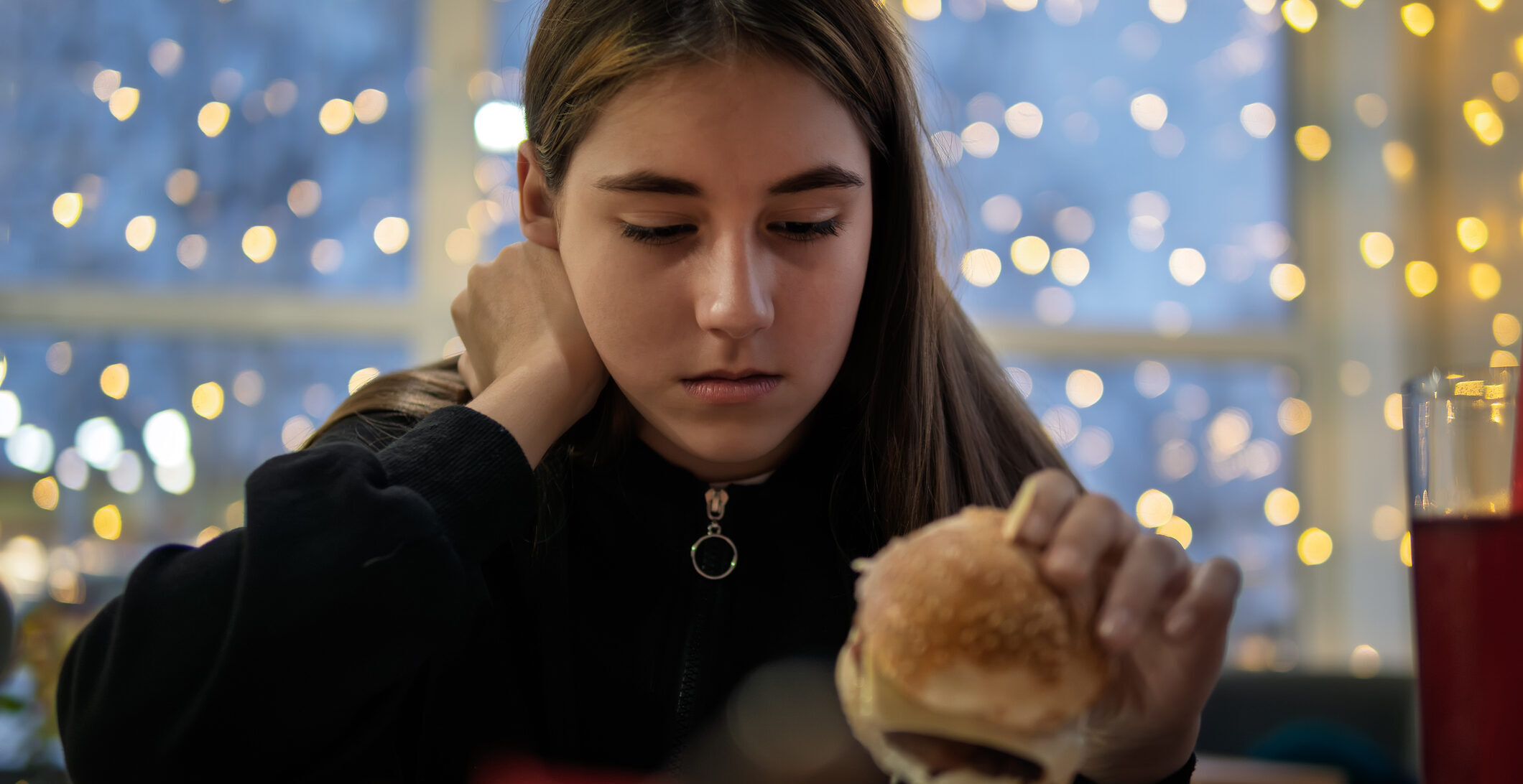 A teenage girl sits at a table with one hand on her neck, holding a cheeseburger on her plate. She looks at it reluctantly.