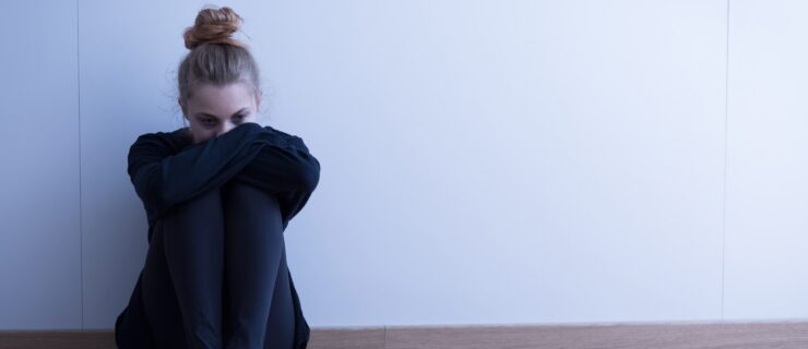 A teenage girl in black leggings, a large black sweater, and sneakers sits on the ground against a wall, hugging her knees to her chest. Her hair is in a messy bun and she looks sad.