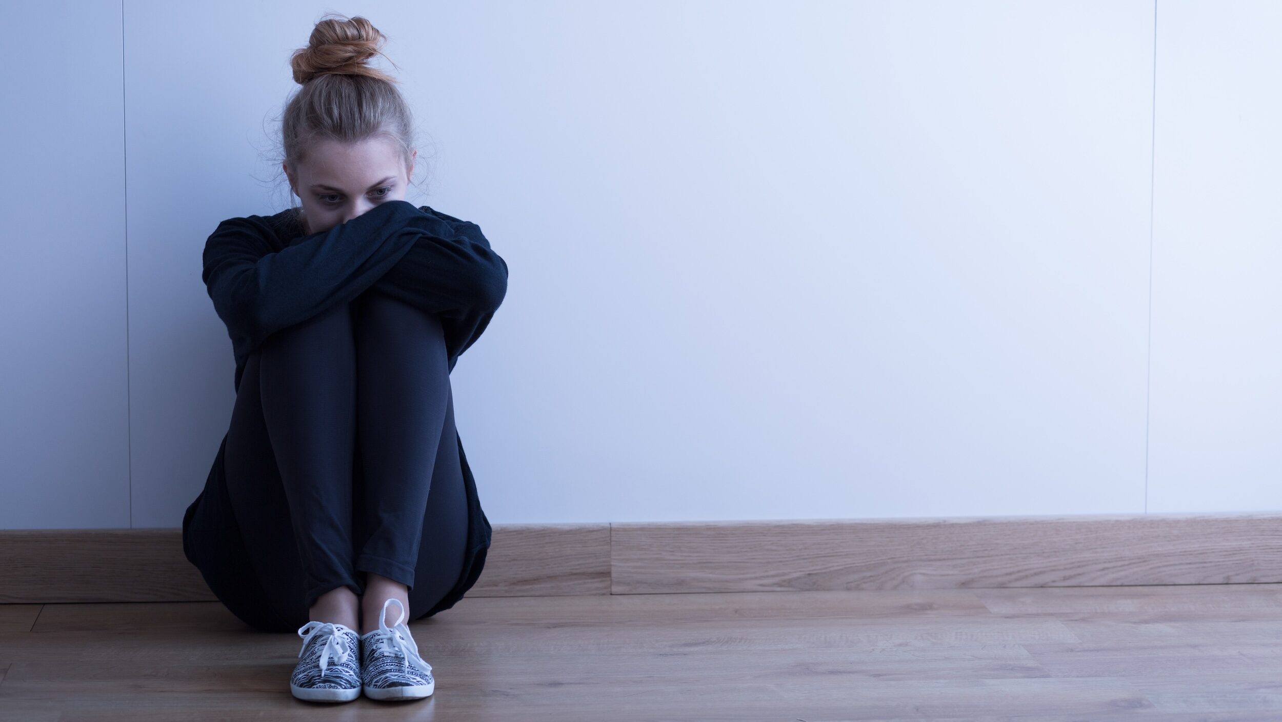 A teenage girl in black leggings, a large black sweater, and sneakers sits on the ground against a wall, hugging her knees to her chest. Her hair is in a messy bun and she looks sad.