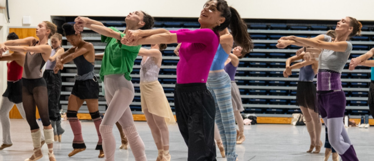 Patrica Delgado, wearing a hot pink half-sleeve leotard with black pants, leads Miami City Ballet dancers in rehearsal. She is shown in the center of the frame, with over ten dancers shown behind her in rows, all wearing colorful layers of dance clothes with pointe or ballet shoes. They all stand facing the left side of the photo, their arms stretched out in front of them, crossed at the forearm with hands loosely clasped together. The dancers look upward or toward their hands; Delgado, smiling, looks from the corner of her eye toward the front.