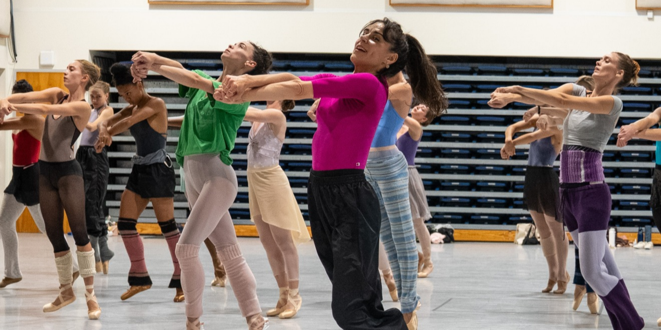 Patrica Delgado, wearing a hot pink half-sleeve leotard with black pants, leads Miami City Ballet dancers in rehearsal. She is shown in the center of the frame, with over ten dancers shown behind her in rows, all wearing colorful layers of dance clothes with pointe or ballet shoes. They all stand facing the left side of the photo, their arms stretched out in front of them, crossed at the forearm with hands loosely clasped together. The dancers look upward or toward their hands; Delgado, smiling, looks from the corner of her eye toward the front.