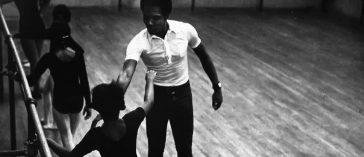 A black-and-white archival photo of Arthur Mitchell, wearing a white t-shirt and dark pants, teaching a class of young ballet students. The dancers face away from the camera and execute a combination with one hand on the barre.