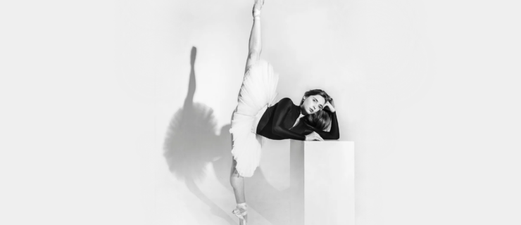 In black-and-white, Hannah Martin is shown in a 180-degree penché en pointe against a white background, casting a defined shadow. She wears a white tutu and a black long-sleeved leotard. Martin casually props her elbow up on a white rectangular box, resting her head on the back of her hand, which she curls into a loose fist. Her other arm rests on the prop in front of her. Her hair is down and she looks calmly into the camera.