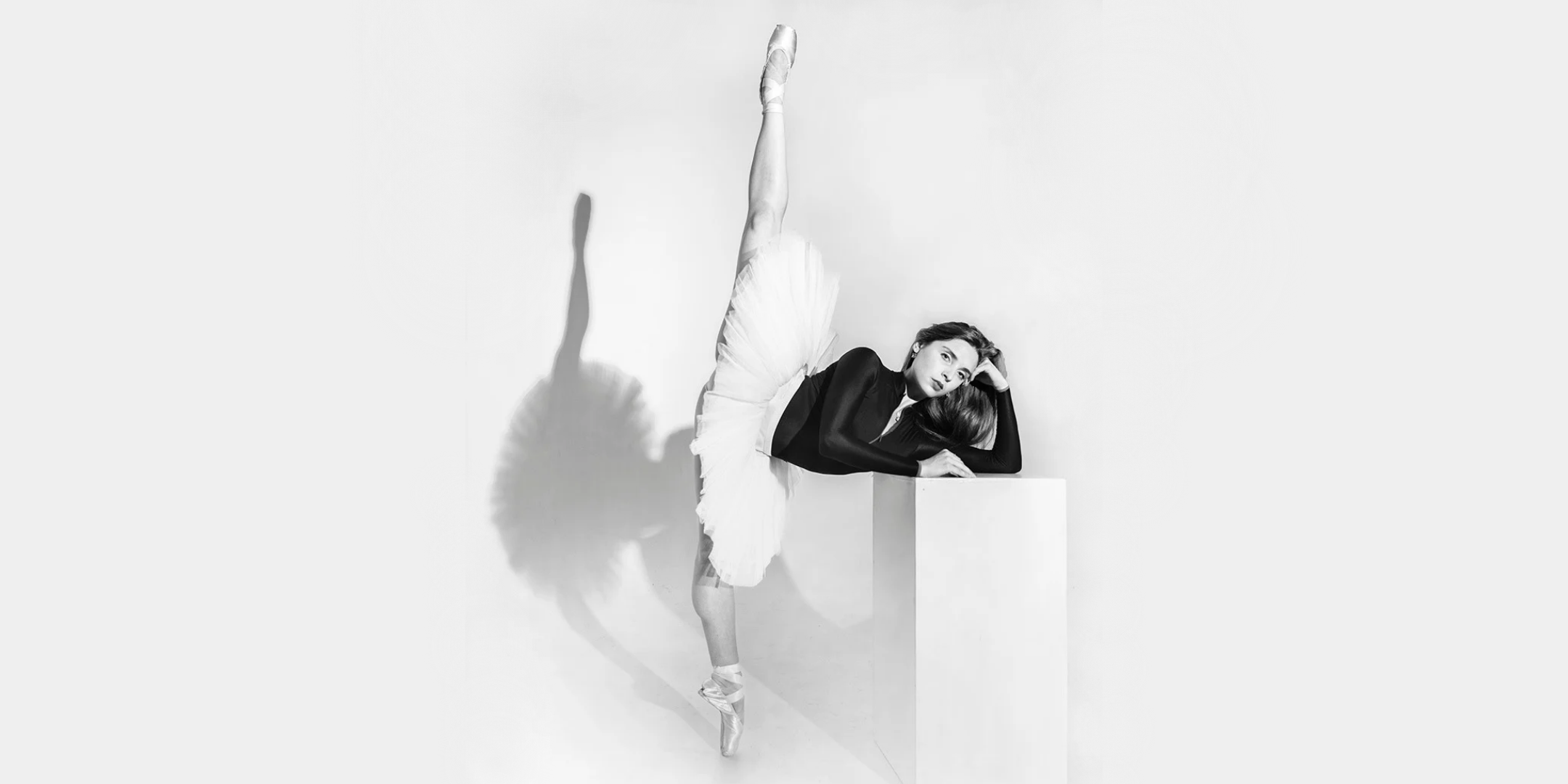 In black-and-white, Hannah Martin is shown in a 180-degree penché en pointe against a white background, casting a defined shadow. She wears a white tutu and a black long-sleeved leotard. Martin casually props her elbow up on a white rectangular box, resting her head on the back of her hand, which she curls into a loose fist. Her other arm rests on the prop in front of her. Her hair is down and she looks calmly into the camera.
