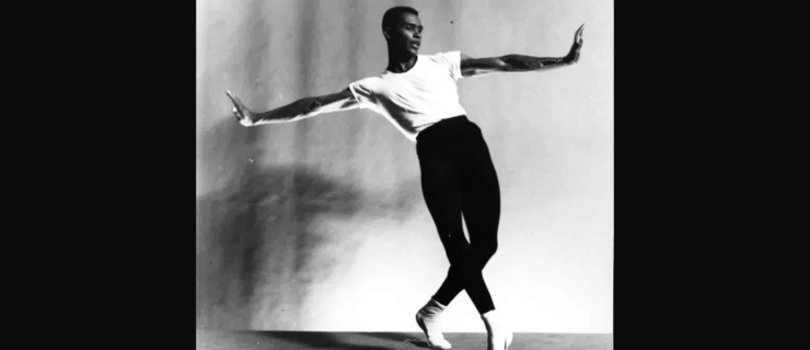 A black-and-white archival image of Arthur Mitchell posing in front of a neutral backdrop, his arms extended to either side with palms flexed up. He leans back with his hips forward, both feet on demi pointe as he crosses his left leg over his right.