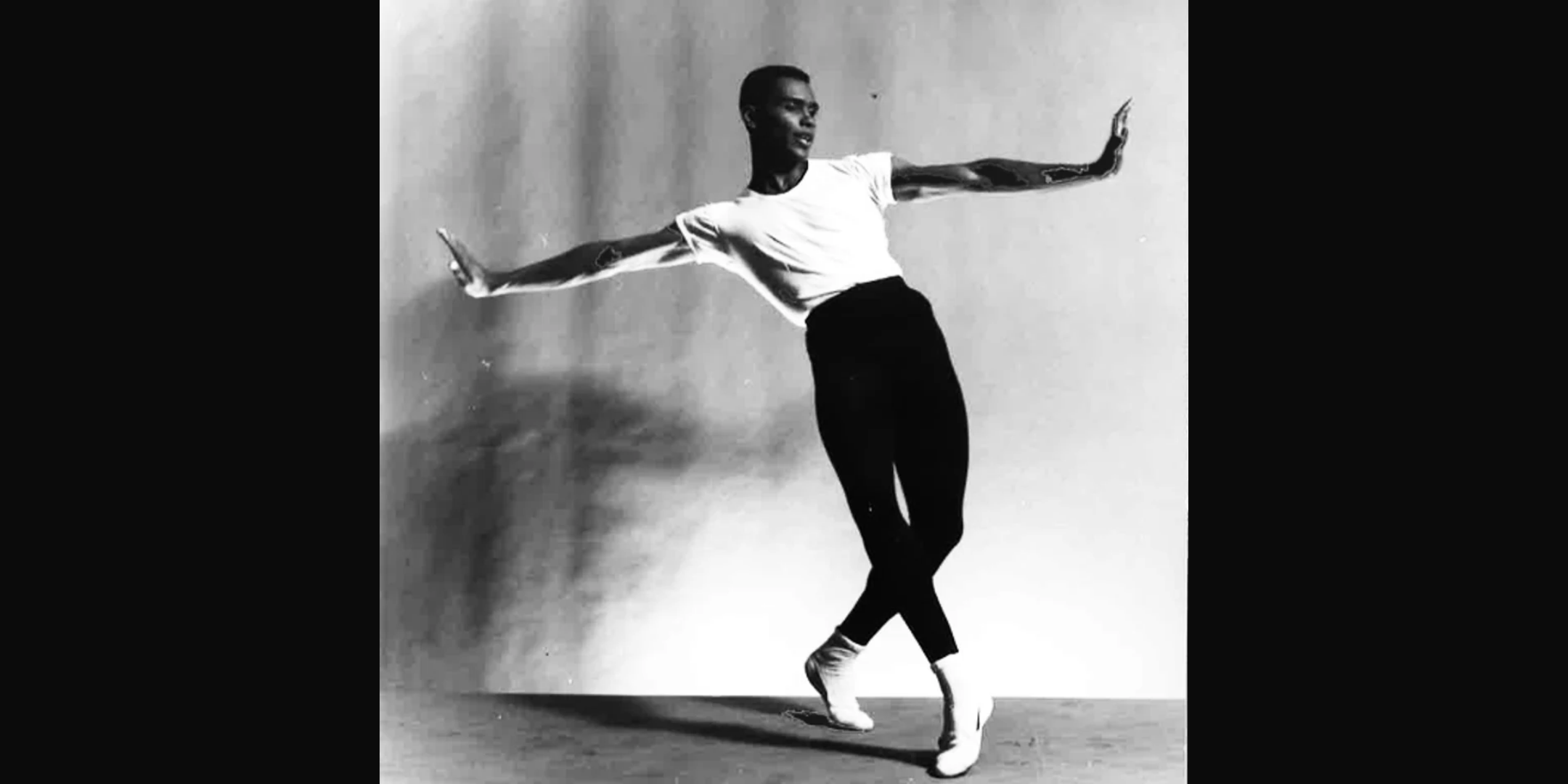 A black-and-white archival image of Arthur Mitchell posing in front of a neutral backdrop, his arms extended to either side with palms flexed up. He leans back with his hips forward, both feet on demi pointe as he crosses his left leg over his right.