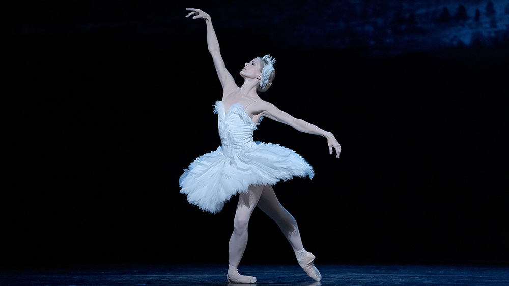Heather Ogden performs as Odette in "Swan Lake," wearing a white classical tutu with feathers and a matching headpiece. She poses in tendu croisé derriere with her front leg in plié, arms extended in a high arabesque position. She looks up and past her top arm with a pained expression.