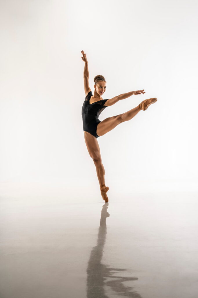 Regan Hutsell poses on pointe with her left leg extended a la seconde, a slight twist in her body as she leans slightly to the right for style. Her arms extend in a high arabesque; she looks over her left fingertips as her arm matches her extended leg's line. She wears a sleeveless black leotard.