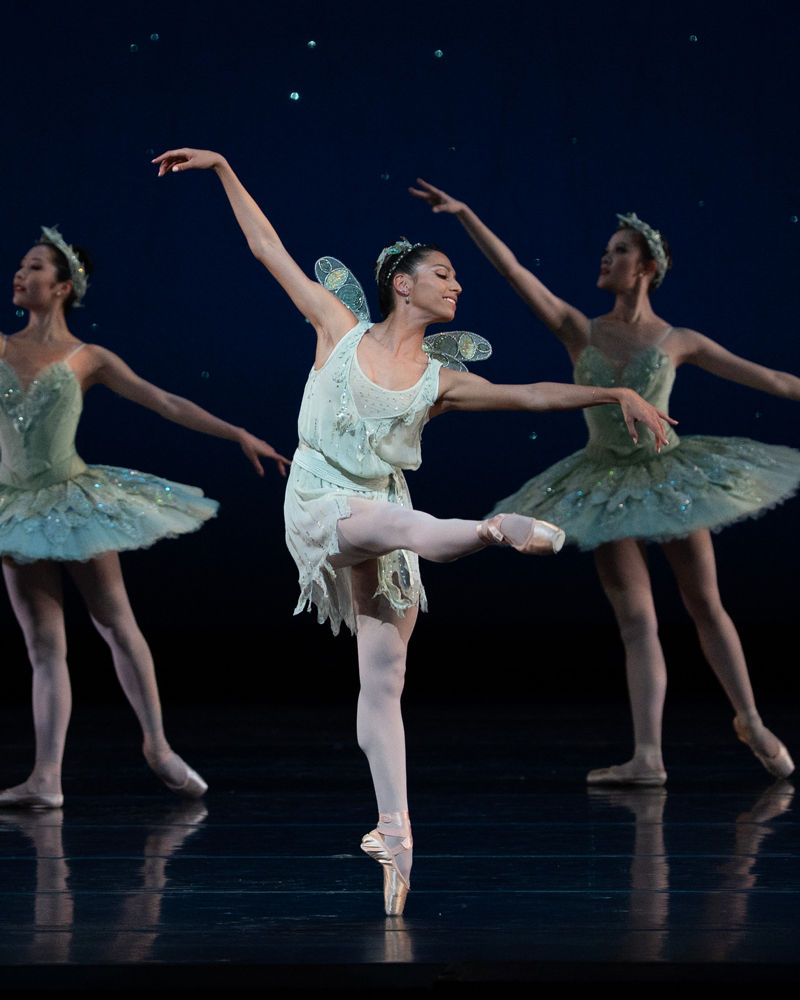 Isabella DeVivo, dressed in a short, light green dress with fairy wings, poses on pointe with one leg croisé devant at 90 degrees. She leans forward slightly with her opposite arm reaching forward, the other extended up and back. Corps dancers in light green tutus stand in tendu back behind her.