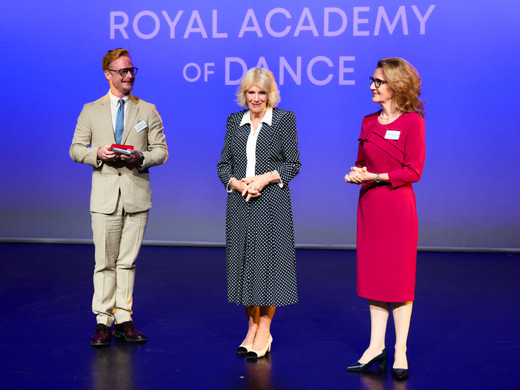 Christopher Wheeldon, Queen Camilla, and Elizabeth Honer stand together on a stage at an awards ceremony, wearing formal attire. Wheeldon holds an awards and smiles at Queen Camilla, who folds her hands and smiles at the audience offscreen. Honer folds her hands, as well, and looks at the queen. Behind them, "Royal Academy of Dance" is projected onto a screen.