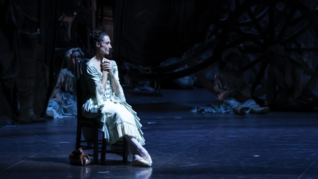 Dorothée Gilbert sits in a chair upstage during a performance of the ballet Manon. She is costumed in an elegant and