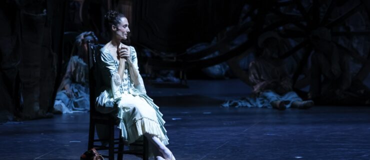 Dorothée Gilbert sits in a chair upstage during a performance of the ballet Manon. She is costumed in an elegant and light-colored ruffled dress and clasps her hands to her chest. She is bathed in cool lighting, and in the background, others in costume sit on the floor.
