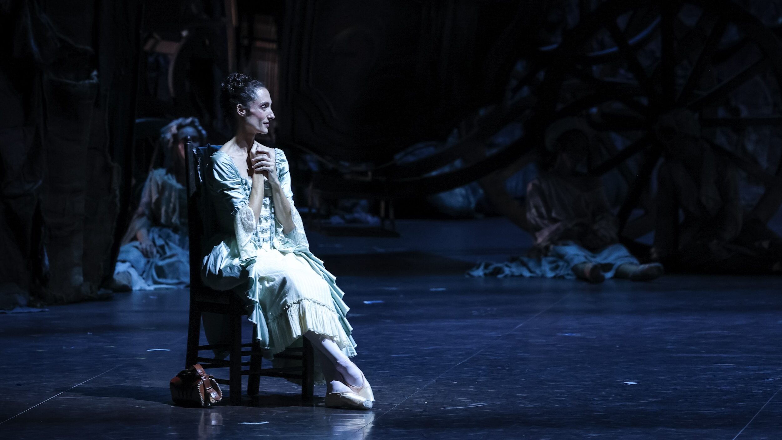 Dorothée Gilbert sits in a chair upstage during a performance of the ballet Manon. She is costumed in an elegant and light-colored ruffled dress and clasps her hands to her chest. She is bathed in cool lighting, and in the background, others in costume sit on the floor.
