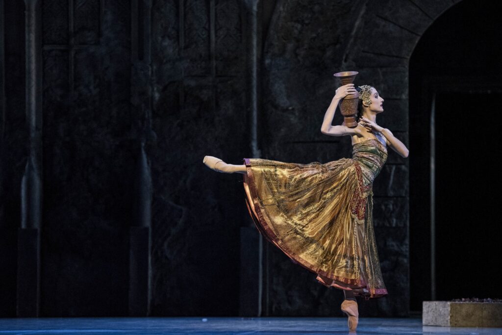 Dorothée Gilbert wears a long, shiny gold dress with red trim and an alaborate grold crown. She does a piqué arabesque on pointe towards stage left, holding a large vase on her right shoulder with both hands.