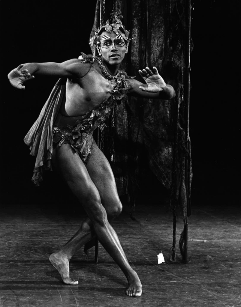 A black-and-white archival photo of Arthur Mitchell dressed as a sprite, performing onstage. He leans his body to the right, arms lifted with one bent, and crosses one leg behind the other as he looks and listens attentively.