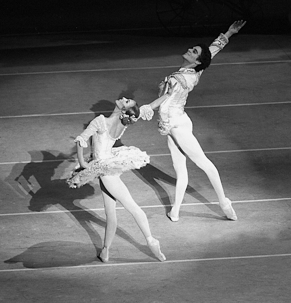 Natalia Makarova and Fernando Bujones perform a pas de deux together onstage. They both stand in tendu derriere towards the upstage right corner, with their left hand on their hip and their right arm up, twisting their upper bodies to their right and throwing their heads back with a flourish. Makarova wears a white tutu and pointe shoes, and Bujones wears white tights and a white bolero jacket,