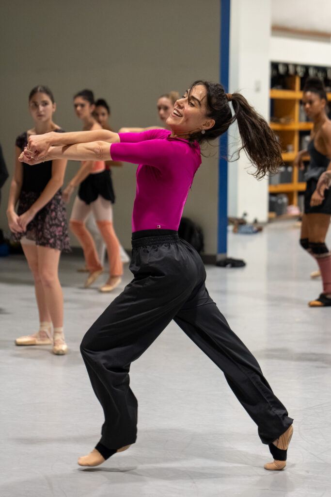 Patricia Delgado demonstrates her choreography for Miami City Ballet dancers in rehearsal. She wears a hot pink half-sleeve leotard with black pants and skin-tone ballet flats. She is captured in motion, her arms stretched out in front of her and crossed at the forearm. She has one foot behind her on a high demi-pointe and the other mid-step in front of her, just above the ground. Delgado leans her head away from her arms and looks slightly to the side, smiling. Dancers in ballet clothes and pointe shoes observe in the background.