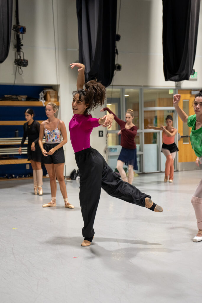 Patricia Delgado is shown demonstrating her choreographing for Miami City Ballet Dancers in rehearsal. She wears a hot pink half-sleeve leotard with black pants and skin-tone pointe shoes. She appears to be landing a jump in a low third