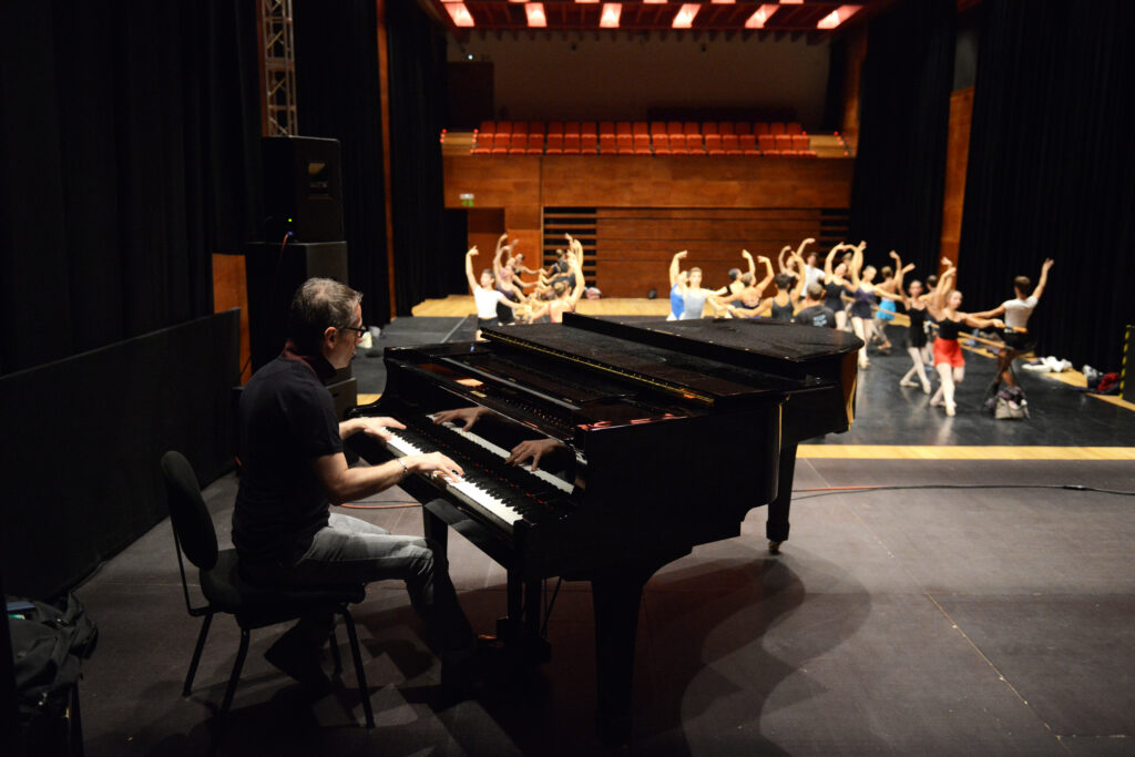 Massimiliano Greco plays on a black piano for a large group of ballet dancers warming up at the barre onstage.