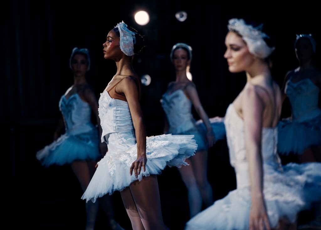 Salomé Tregre is shown in profile onstage during a performance of Swan Lake. She wears a white tutu and feathered headpiece and stands in b plus with her arms in demi seconde. Other dancers in the same costume pose in a line in the background and one is shown in the foreground slightly out of focus.