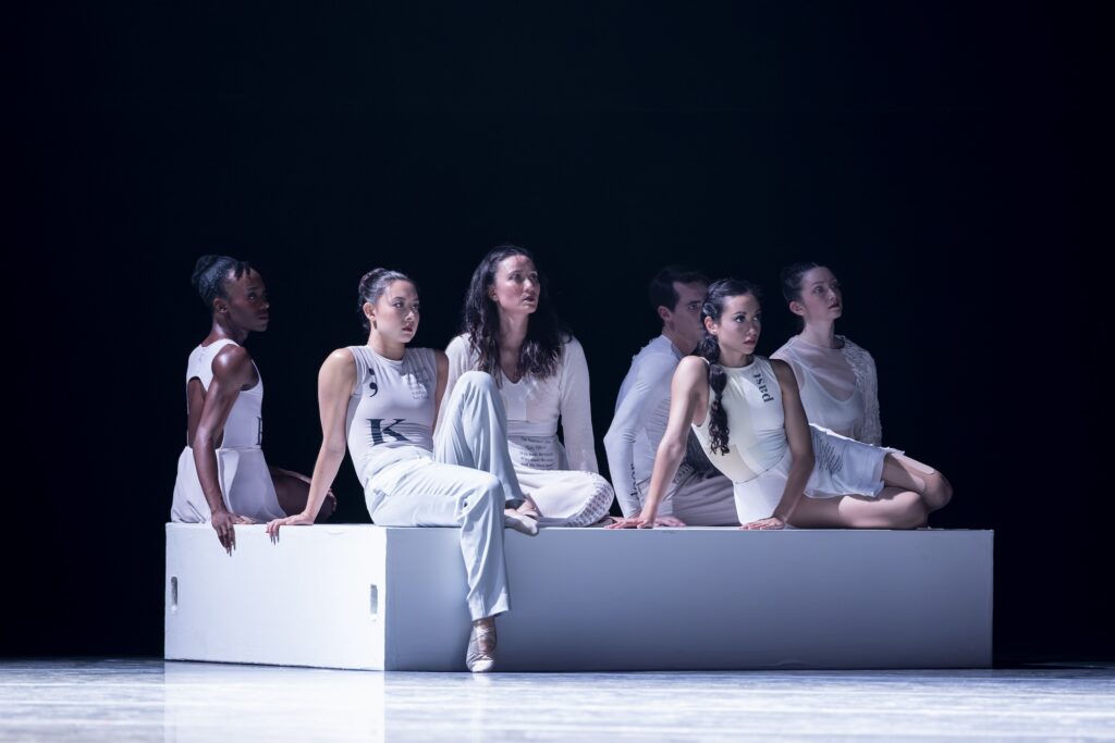 A group of dancers in white costumes sit on a white, rectangular box on a darkened stage. They looks towards something off camera and downstage right.