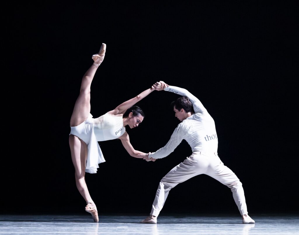 Sarah Gabrielle, wearing a white leotard and short ballet skirt, holds hands with her dance partner, Mark Cuddifee, who is costumed in white pants and a long-sleeved shirt. Ryan tilts towards her left on pointe, her right leg lifted high to create a split, as Cuddifee lpliés in second position, pulling her right arm.