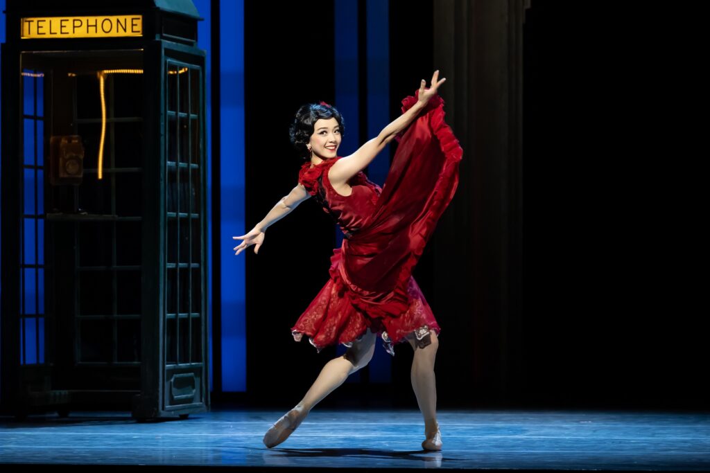 Sirui Liu, wearing a dark red knee-length dress, is shown in profile during a performance onstage. She lunges out towards stage left, lifting her right arm pu and left arm back, and looks out towards the audience with a smile. The train of her skirt flies up towards her right hand. A telephone booth is shown in the background.