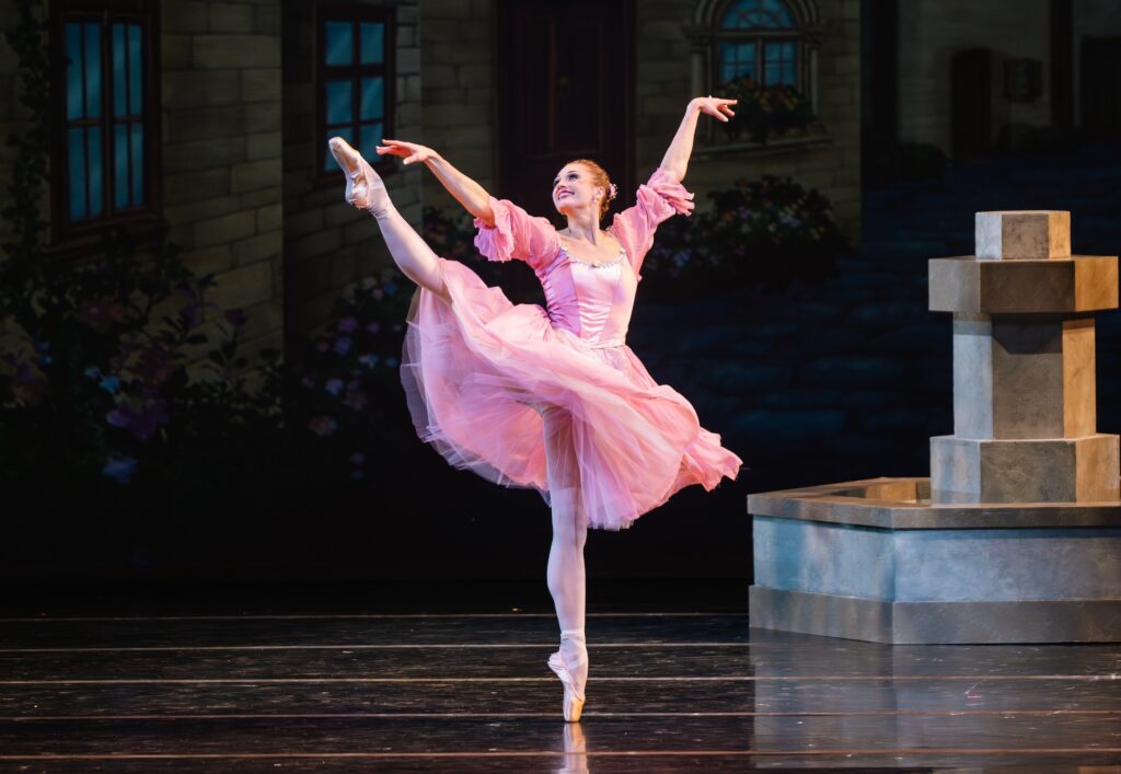 Madeline rogers, wearing a pink dress, tights, and pointe shoes, does a déveleoppé à la seconde onstage with her right leg. She lifts her arms up in a V shape and looks out over her right shoulder with a large smile. Her red hair is pulled back in a bun.