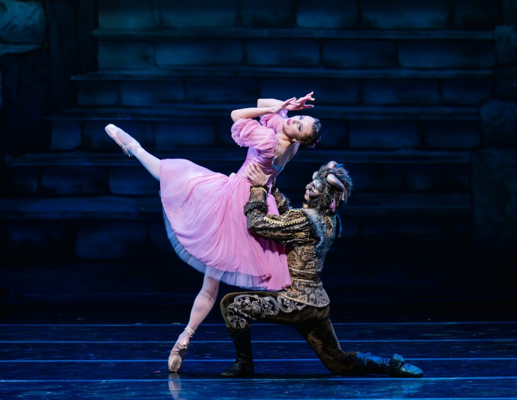 Two dancers perform a pas de deux. The woman, wearing a pink dress, pink tights, and pointe shoes, stands on her right leg on pointe and extends her left leg in front. The man, wearing a beast costume and gold and black tunic and pants, kneels behind the ballerina and holds her by the waist as she leans back and covers her face with her hands.
