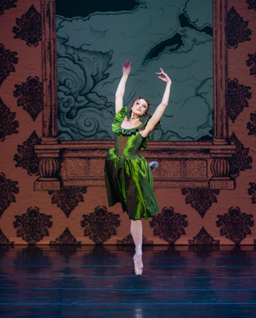 Madelien Rogers, costumed in a bight green satin dress, jumps up onstage with her arms in high fifth and her right leg behind her in attitude. Her red hair is loosely pulled back.