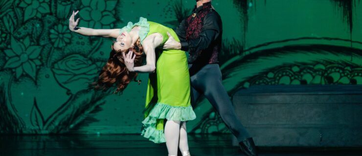 Madeline Rogers, wearing a bright green knee-length dress, stands in sous-sus on pointe facing Kiefer Curtis and bends her upper body back with her arms up as Curtis holds her by the waist. Her long red hair flows behind her. Curtis is costumed as Dracula, wearing a greay wig, black tights, boots, and ashirt and a dark red vest. He opens his mouth menacingly, showing his fangs.