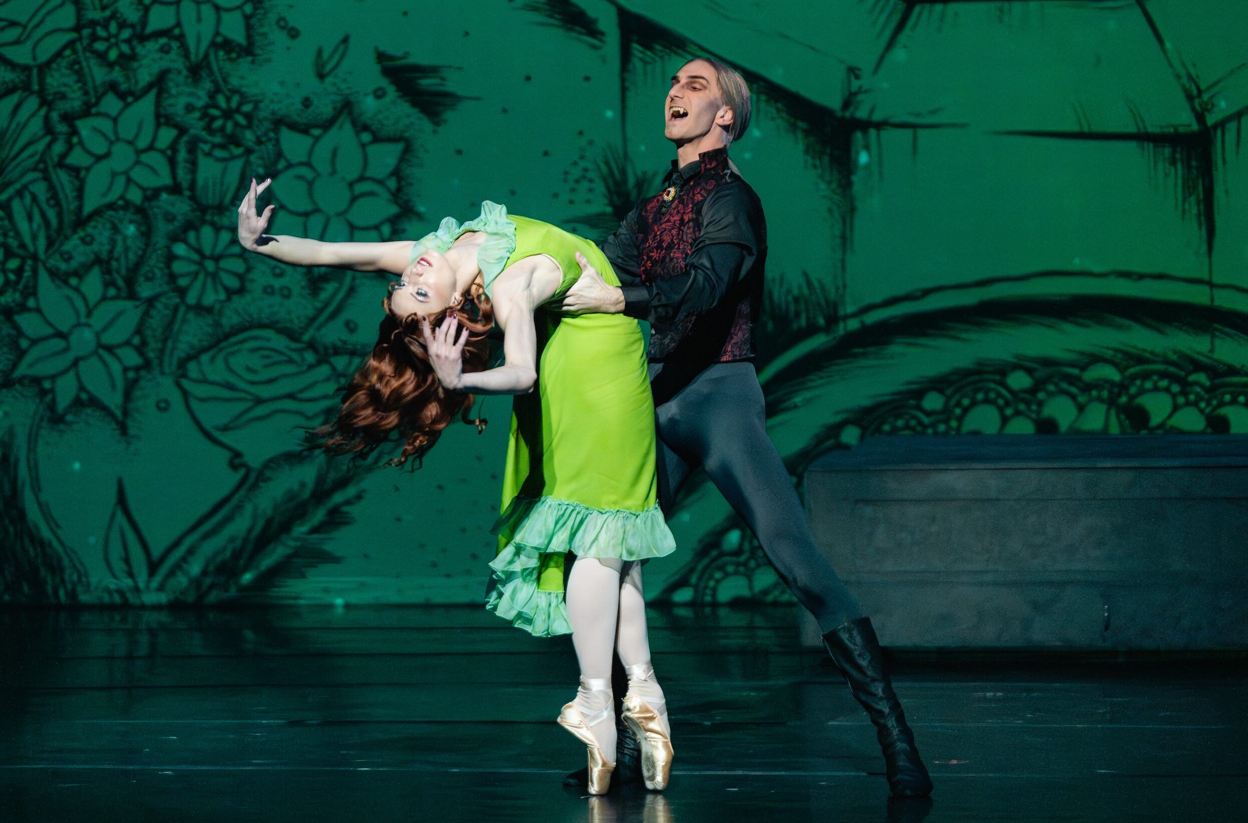 Madeline Rogers, wearing a bright green knee-length dress, stands in sous-sus on pointe facing Kiefer Curtis and bends her upper body back with her arms up as Curtis holds her by the waist. Her long red hair flows behind her. Curtis is costumed as Dracula, wearing a greay wig, black tights, boots, and ashirt and a dark red vest. He opens his mouth menacingly, showing his fangs.