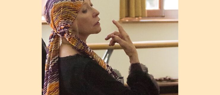 Natalia Makarova is shown in profile from the ribs up in a ballet studio. She wears a colorful headscarf and long-sleeved black sweater, and gracefully raises her right hand up towards her chin as she looks down and out to her right.