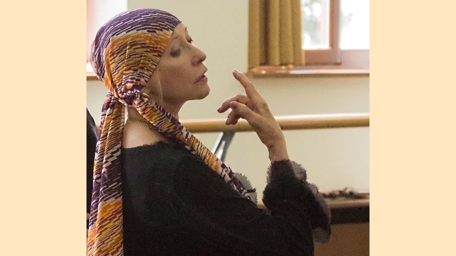 Natalia Makarova is shown in profile from the ribs up in a ballet studio. She wears a colorful headscarf and long-sleeved black sweater, and gracefully raises her right hand up towards her chin as she looks down and out to her right.