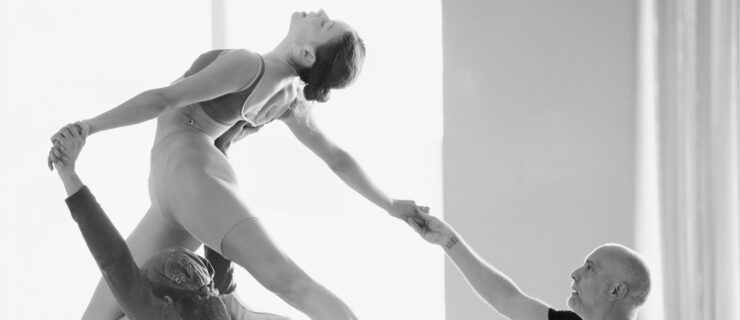 In grayscale: Isabella Kinch, Larkin Miller, and David Dawson are shown rehearsing a lift. Kinch, wearing a tank leotard with pointe shoes and tights cut at mid thigh, kneels on Miller's shoulder, one leg extended behind her in an arabesque. She arches backward, her face turned toward the ceiling in profile. Miller, wearing a long-sleeved shirt and a bandana, supports her with his arms above his head - one holding her back and the other holding one of her hands. Dawson, wearing a black short-sleeved shirt, stands behind the duo, holding Kinch's other outstretched hand and looking up toward her.