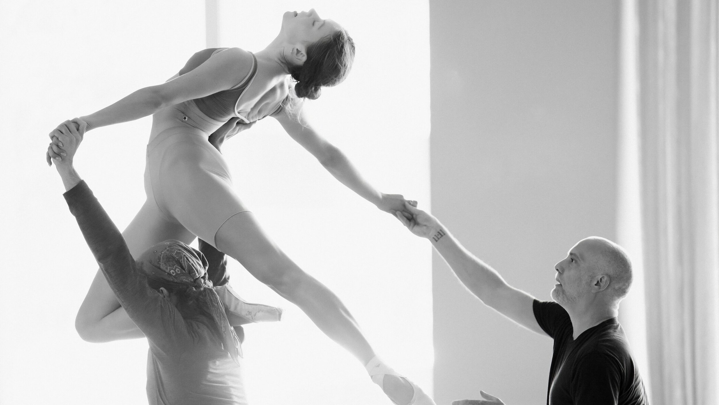 In grayscale: Isabella Kinch, Larkin Miller, and David Dawson are shown rehearsing a lift. Kinch, wearing a tank leotard with pointe shoes and tights cut at mid thigh, kneels on Miller's shoulder, one leg extended behind her in an arabesque. She arches backward, her face turned toward the ceiling in profile. Miller, wearing a long-sleeved shirt and a bandana, supports her with his arms above his head - one holding her back and the other holding one of her hands. Dawson, wearing a black short-sleeved shirt, stands behind the duo, holding Kinch's other outstretched hand and looking up toward her.