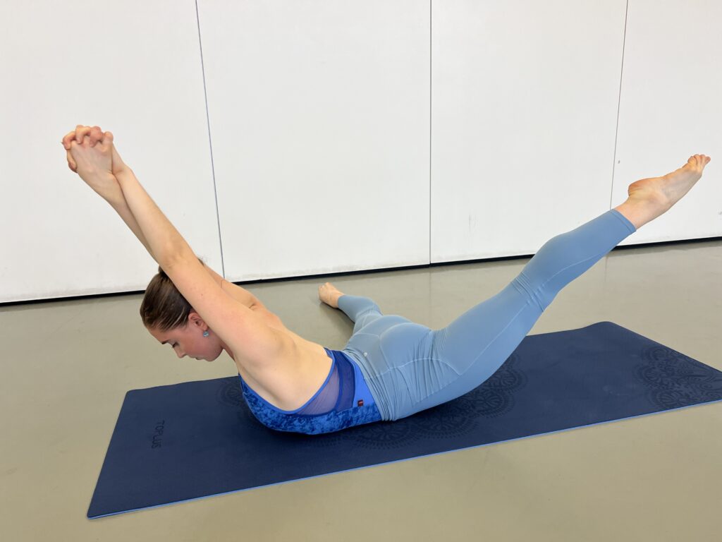 Hannah Martin demonstrates an exercise on a navy yoga mat. She is laying prone, one leg extended down away from her body and the other at a ninety-degree angle to her side. Her arms are stretched above and behind her head, crossed at the forearms with hands clasped. She arches her upper torso off the floor and lifts her bottom leg at around a forty-five degree angle. She keeps the leg to the side flat on the floor. She wears a bright blue halter leotard and light blue leggings.