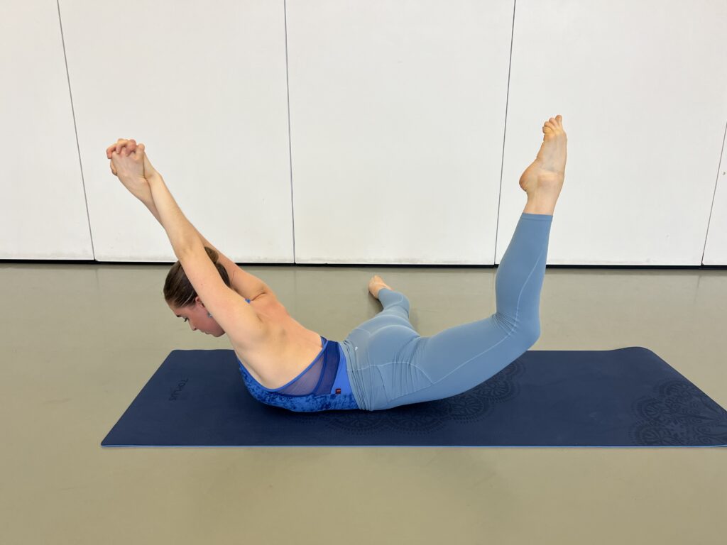 Hannah Martin demonstrates an exercise on a navy yoga mat. She is laying prone, one leg extended down away from her body and the other at a ninety-degree angle to her side. Her arms are stretched above and behind her head, crossed at the forearms with hands clasped. She arches her upper torso off the floor and lifts her bottom leg off the floor, her thigh at a forty-five-degree angle to the floor and her knee bent, pointing her toes to the ceiling. Her other leg remains flat to the ground beside her. She wears a bright blue halter leotard and light blue leggings.